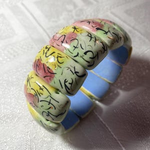 Könnte beinhalten: Ein farbenfrohes Stretch-Armband mit rechteckigen Gliedern. Die Glieder sind mit gelben, rosa und grünen Blumenmustern und schwarzen Akzenten im Kalligraphie-Stil bemalt. Die Unterseite der Glieder ist hellblau.