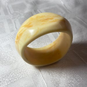 May include: A chunky, ivory-coloured bangle bracelet with a marbled, amber-toned pattern. The smooth, rounded surface reflects light, highlighting the warm hues. This vintage-style bracelet is a statement piece.