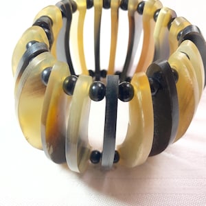 Könnte beinhalten: Ein elastisches Armband mit abwechselnd gebogenen Segmenten in Gelb, Schwarz und Grau. Kleine schwarze Perlen verbinden die Segmente. Das Armband hat eine natürliche, organische Ästhetik.