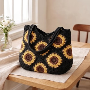 May include: A black crochet tote bag with a sunflower design. The sunflowers are made with yellow and brown yarn, set against a black background. The bag has two black handles. The bag is on a wooden table.