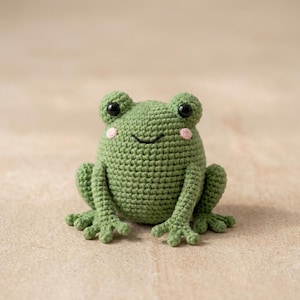 May include: A handmade green crochet frog with black eyes and pink cheeks. The frog is sitting upright with its arms and legs extended. The frog is a cute, whimsical toy.