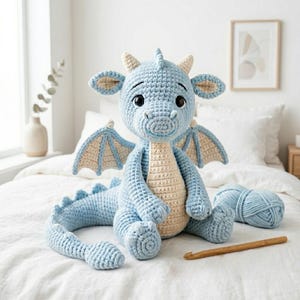 May include: A light blue and cream colored crocheted dragon plush toy with large dark eyes, small horns, and wings. The dragon has a long tail with spikes and is positioned on a white surface next to a ball of blue yarn and a wooden crochet hook.