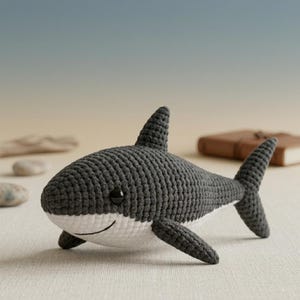 Puede incluir: Un juguete de tiburón tejido a ganchillo en tonos grises y blanco. El tiburón tiene una cara sonriente, ojos negros y una aleta dorsal. El juguete está hecho de hilo suave y está diseñado para ser un compañero tierno.