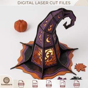 May include: A purple and black laser-cut wooden witch hat decoration featuring cutouts of a witch on a broom, crescent moons, stars, bats, and flames. The structure is illuminated from within and accompanied by a small orange pumpkin and leaf.