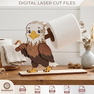 May include: A laser-cut wooden eagle toilet paper holder. The eagle, with brown, tan, and white details, holds a roll of white toilet paper. The base is a light brown rectangle. The text "DIGITAL LASER CUT FILES" is at the top.