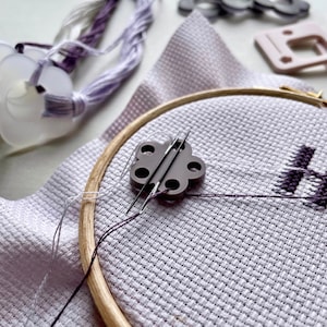 Könnte beinhalten: Nahaufnahme eines violetten Stickrahmens mit einem blumenförmigen Nadelfadenzieher. Der Fadenzieher hat sechs Löcher für Nadeln und ist aus Kunststoff. Der Rahmen befindet sich auf einem weißen Stoffhintergrund.