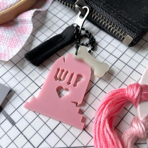 Puede incluir: Un dije rosa en forma de lápida con las letras "WIP" recortadas, junto con una forma de corazón. Un dije blanco en forma de hueso y un tirador de cremallera negro están adjuntos. Los artículos están sobre un fondo de cuadrícula.