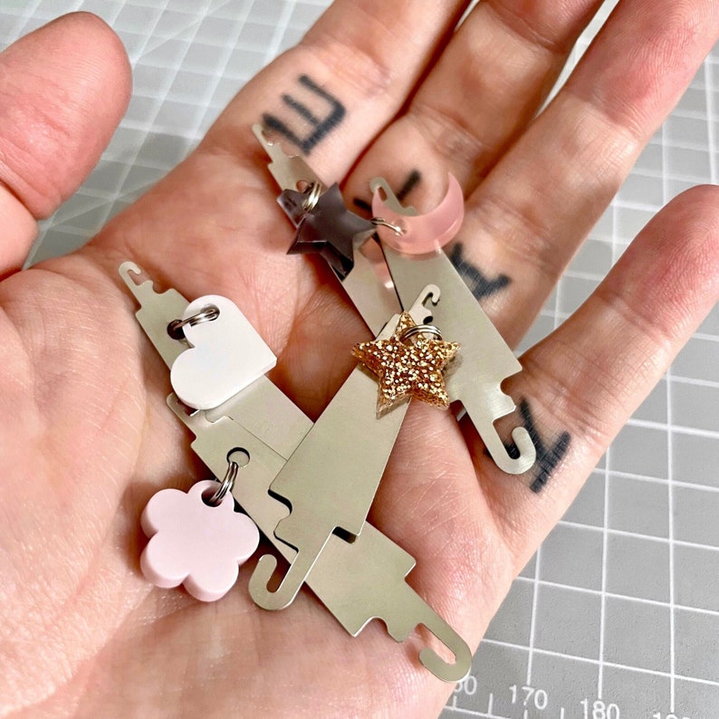 May include: A set of four silver metal stitch markers with different colored charms. The charms include a pink flower, a white heart, a gold star, and a pink crescent moon.