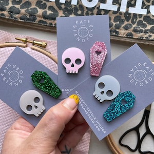 Op de afbeelding: Drie sets magnetische naaldmagneten op grijze kaarten, elk met een schedel en een doodskist. De schedels zijn lichtroze, lichtgrijs en wit. De doodskisten zijn glinsterend roze, groen en blauw, elk met een kleine hartvormige uitsparing.