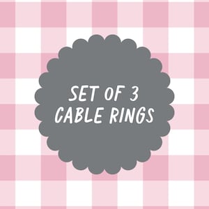 Op de afbeelding: Een set van drie grijze kabelringen op een roze en witte ruitjesachtergrond. De tekst "SET OF 3 CABLE RINGS" staat in wit op de ringen.