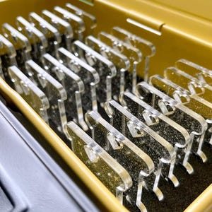 May include: A close-up shot of a yellow container filled with multiple clear, acrylic, comb-shaped objects. The objects are arranged neatly inside the container, which has a black interior. The container is open, revealing the contents.