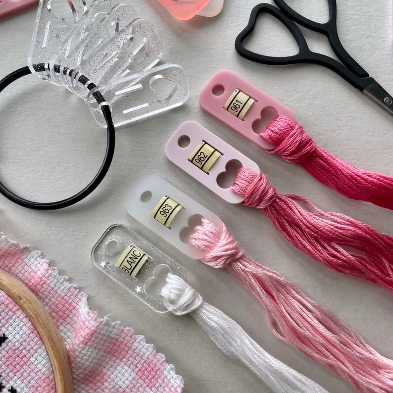 May include: Four plastic thread organizers with pink and white thread spools. The organizers have a heart-shaped opening and a small label with a number on it. The numbers on the labels are 961, 962, 963, and BLANC.