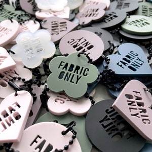 Pode incluir: Uma variedade de etiquetas de tecido coloridas com o texto "FABRIC ONLY" em letras pretas. As etiquetas têm formas diferentes, incluindo corações, flores e círculos. As etiquetas estão presas a correntes pretas.