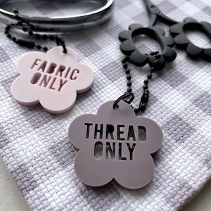 Puede incluir: Primer plano de dos etiquetas con forma de flor con las palabras "FABRIC ONLY" y "THREAD ONLY" en negrita. Las etiquetas están unidas a cadenas de bolas negras. También se ven unas tijeras negras y un dedal plateado.