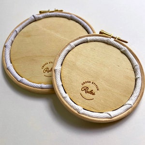 May include: Two wooden embroidery hoops with white fabric lining. The hoops have a brown wood finish and a gold screw. The text "Cross Stitch Rules" is printed on the bottom hoop.