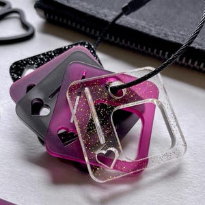 May include: A collection of square luggage tags in black, purple, and clear. Each tag features a heart-shaped cutout and is attached to a black wire loop. The clear tag is speckled with gold glitter.
