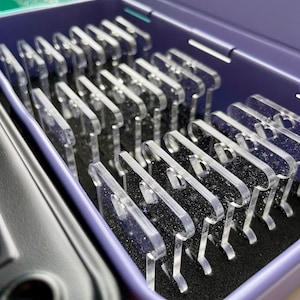 May include: A purple box containing rows of clear, acrylic shapes with star-like speckles. The shapes have a hook design. The box is open, revealing the contents, and is next to a black box.