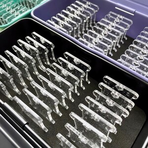 May include: Clear acrylic display stands arranged in open containers. The stands are rectangular with a small heart-shaped cutout. The containers are in shades of green, purple, and black, with a black foam base.
