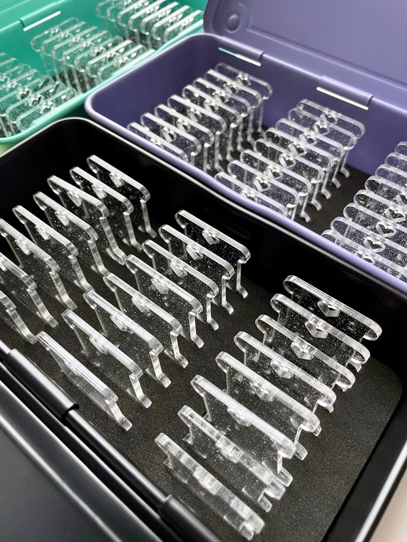 May include: Clear acrylic display stands arranged in open containers. The stands are rectangular with a small heart-shaped cutout. The containers are in shades of green, purple, and black, with a black foam base.