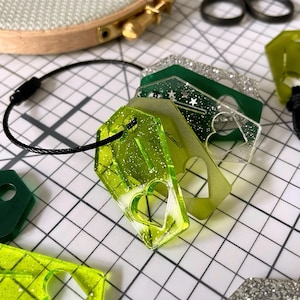 May include: A selection of green and clear acrylic tags with heart and star cutouts, joined by a black cord. Some tags have glitter accents. The tags are on a white grid surface, with a wooden embroidery hoop in the background.