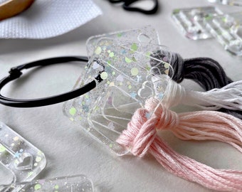 May include: Close-up of a craft project setup. Clear, glitter-filled thread organizers with heart-shaped cutouts are attached to a black ring. Pink, white, and gray embroidery floss are attached. Black scissors and a white fabric are in the background.