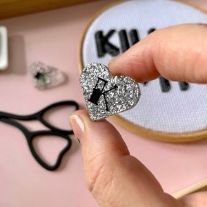 May include: A silver glitter heart-shaped pin with a black graphic of a sewing machine. The pin is held between fingers. Other craft supplies are in the background, including scissors and a small embroidery hoop.