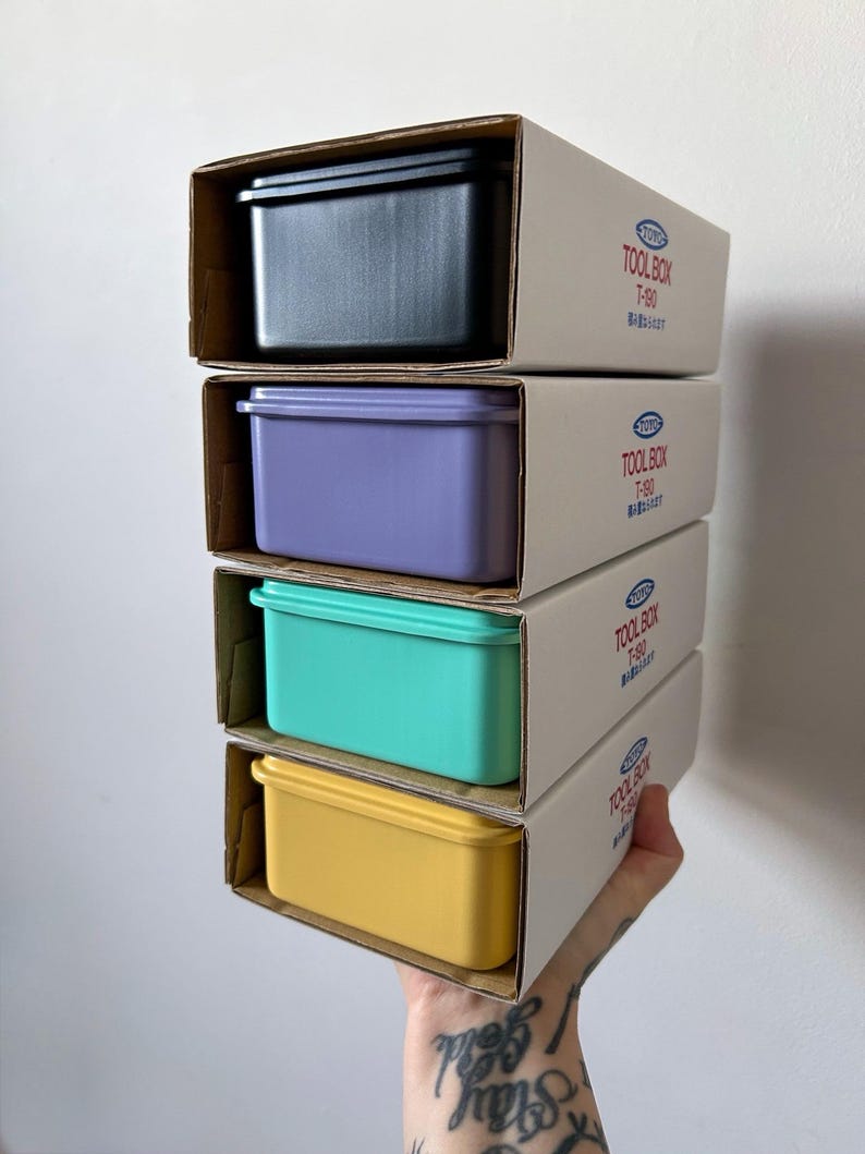 May include: A stack of four colorful plastic toolboxes in their cardboard packaging. The boxes are black, purple, teal, and yellow. The boxes are labeled "TOOLBOX T-80".