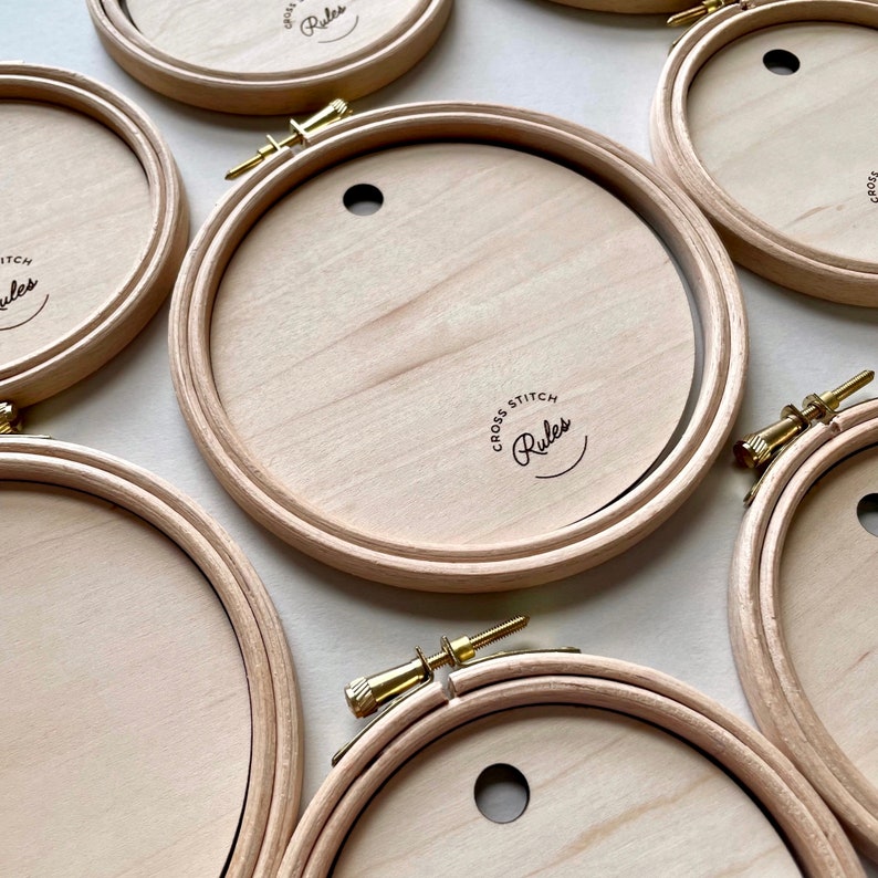 May include: Five wooden embroidery hoops with a gold screw closure. The hoops are all the same size and have the words "Cross Stitch Rules" printed on them.