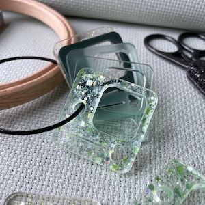 May include: A stack of clear, square phone cases with glitter and green accents. The cases have a heart-shaped hole and are attached to a black cord. Other craft supplies, including scissors and an embroidery hoop, are in the background.