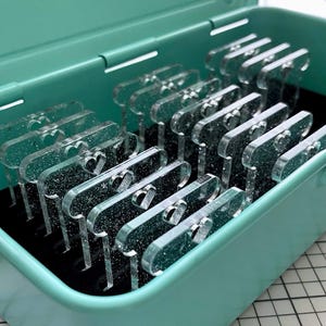 May include: A collection of clear acrylic heart-shaped charms with glitter, neatly arranged inside a teal storage box. The charms have a heart cutout design. The box is open, revealing the contents.