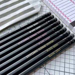 May include: A set of nine black pencils with erasers, arranged in rows. The pencils feature printed text in white, pink, and gold foil, including the phrases STITCHERS GONNA STITCH, FLOSS BOSS, and SEAMS LEGIT, repeated across the different colours.