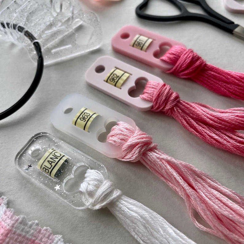 May include: Four clear plastic thread organizers with pink and white embroidery floss. The organizers have a heart-shaped opening and a label with the numbers 961, 962, 963, and BLANC.