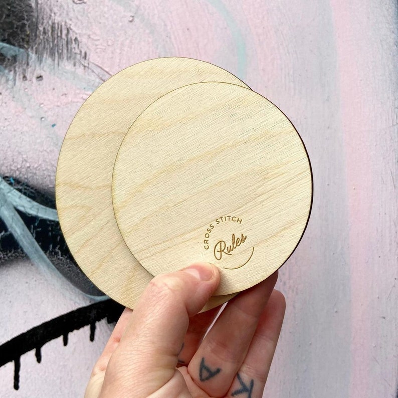 May include: Two round wooden embroidery hoops, one nested inside the other. The smaller hoop has the text "Cross Stitch Rules" engraved on it.