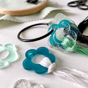 May include: A set of turquoise and clear plastic flower-shaped thread organisers with white and green embroidery floss. The organisers are attached to a black ring.