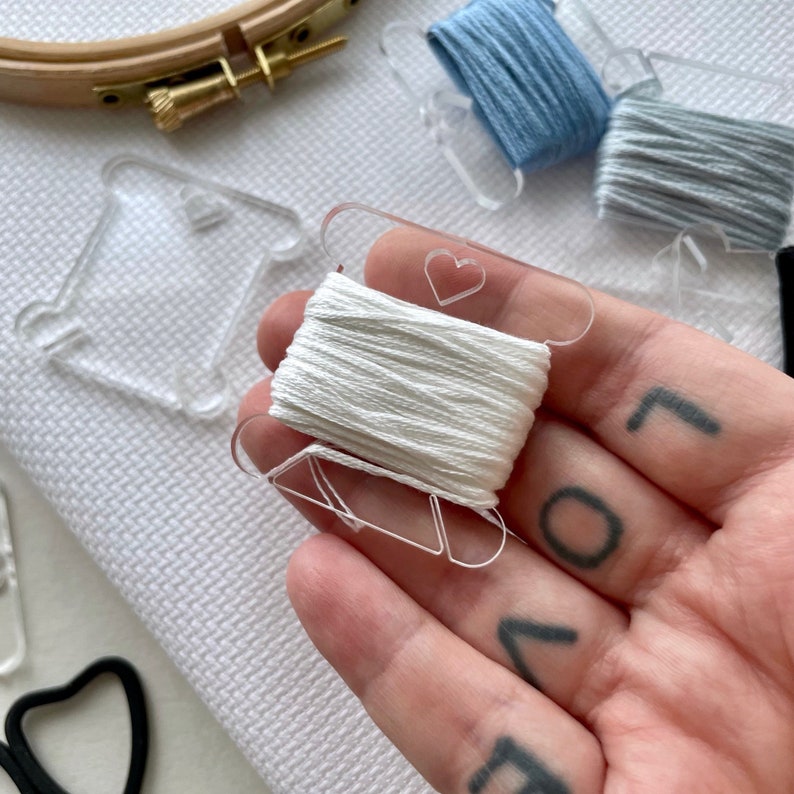 May include: A close-up of white embroidery floss on a clear plastic bobbin with a heart-shaped cutout. Other bobbins with blue and gray floss are visible, along with a wooden embroidery hoop and scissors. The background is white fabric.