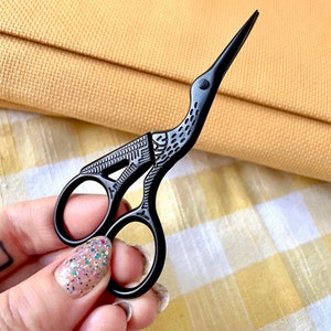 May include: Black crane-shaped embroidery scissors. The scissors have a detailed design on the handles and a sharp, pointed tip. The scissors are held against a yellow and white checkered background.
