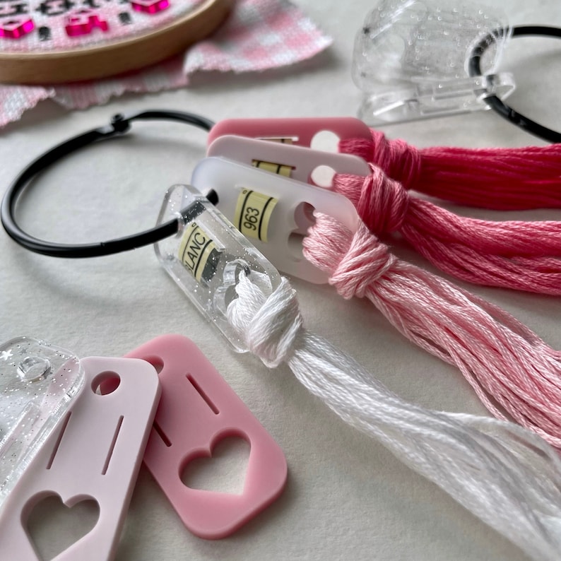 May include: A set of pink and white embroidery floss spools with a black ring and clear plastic holders. The spools are labeled with the number "963" and the word "DMC".