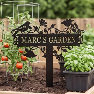 May include: A black metal garden sign featuring the text MARC'S GARDEN in a serif font, decorated with cut-out silhouettes of a bird, a butterfly, and floral vines, mounted on a stake positioned in soil between a tomato plant and a potted herb.