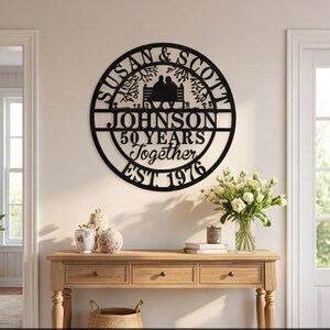 以下が含まれることがあります： 「Susan & Scott」「Johnson」「50 Years Together」「EST 1976」の文字が入った黒い金属製の壁掛けアート。円形のデザインで、ベンチに座るカップルのシルエットと木の枝が描かれています。