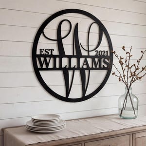 Puede incluir: Un letrero de pared circular de metal negro que presenta una letra W grande y estilizada en el centro. Una barra horizontal en el medio muestra el texto EST 2021 y el nombre WILLIAMS en letras mayúsculas, integrado en el marco circular.