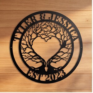 Puede incluir: Un letrero de pared circular de metal negro con un diseño de árbol cortado con láser cuyas ramas forman una forma de corazón en el centro. El texto TYLER & JESSICA está cortado en el borde superior y EST 2023 en el borde inferior.