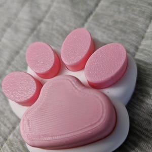 May include: A close-up of a paw-shaped badge reel. The badge reel features a white base with a pink paw print design. The paw print has five pink pads. The badge reel has a metal clip and a retractable cord.
