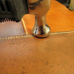 Peut inclure: Gros plan sur un travail du cuir en cours. Un marteau argent&eacute; est plac&eacute; au-dessus d'une couture sur du cuir marron et beige. Le cuir a un aspect textur&eacute; et la couture est r&eacute;alis&eacute;e avec un fil jaune clair. Le manche du marteau est noir.