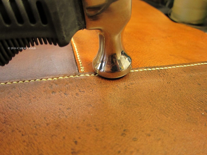 Peut inclure: Gros plan sur un travail du cuir en cours. Un marteau argent&eacute; est plac&eacute; au-dessus d'une couture sur du cuir marron et beige. Le cuir a un aspect textur&eacute; et la couture est r&eacute;alis&eacute;e avec un fil jaune clair. Le manche du marteau est noir.