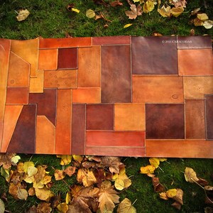 Peut inclure: Un article rectangulaire en patchwork de cuir dans les tons marron, orange et beige. Le motif est compos&eacute; de diverses formes g&eacute;om&eacute;triques cousues ensemble. Le texte "&copy; 2016 CARACODDA" est visible dans le coin sup&eacute;rieur droit. L'article est pr&eacute;sent&eacute; sur un lit d'herbe verte et de feuilles mortes.