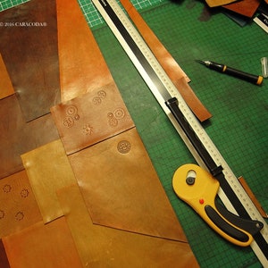 Peut inclure: Un espace de travail artisanal avec diverses pi&egrave;ces de cuir marron de diff&eacute;rentes formes et nuances. Un cutter rotatif jaune, une r&egrave;gle m&eacute;tallique et un cutter sont sur un tapis de coupe vert. Certaines pi&egrave;ces de cuir ont des motifs estamp&eacute;s.
