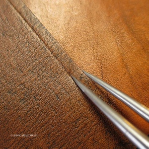 Peut inclure: Gros plan de deux al&egrave;nes en m&eacute;tal argent&eacute; pointant vers une couture sur un morceau de cuir marron. Le cuir a une surface textur&eacute;e et une couleur marron plus fonc&eacute;e au niveau de la couture. L'image est bien &eacute;clair&eacute;e, mettant en valeur les d&eacute;tails du cuir et des outils.