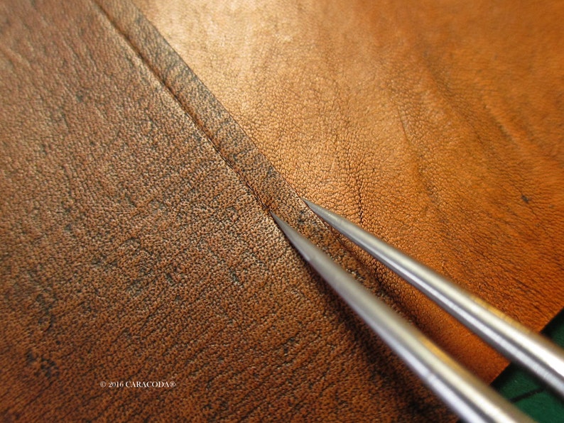 Peut inclure: Gros plan de deux al&egrave;nes en m&eacute;tal argent&eacute; pointant vers une couture sur un morceau de cuir marron. Le cuir a une surface textur&eacute;e et une couleur marron plus fonc&eacute;e au niveau de la couture. L'image est bien &eacute;clair&eacute;e, mettant en valeur les d&eacute;tails du cuir et des outils.