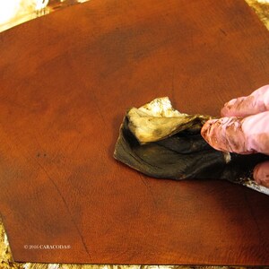 Peut inclure: Gros plan sur une pi&egrave;ce de cuir brun rouge&acirc;tre en cours de teinture. Une main gant&eacute;e applique une teinture fonc&eacute;e avec un chiffon, soulignant la texture et les variations de couleur du cuir. L'image met l'accent sur le traitement du cuir.