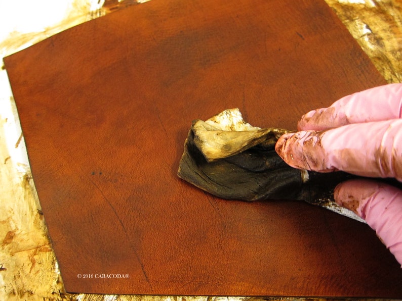 Peut inclure: Gros plan sur une pi&egrave;ce de cuir brun rouge&acirc;tre en cours de teinture. Une main gant&eacute;e applique une teinture fonc&eacute;e avec un chiffon, soulignant la texture et les variations de couleur du cuir. L'image met l'accent sur le traitement du cuir.